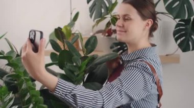 Çekici neşeli beyaz kadın çiçekçi akıllı telefon kullanarak çiçekçide bitkilerin fotoğrafını çekiyor. Gülümseyen neşeli, güzel, yetişkin bir bayan çalışırken eğleniyor. Bahçıvanlık ve hobi kavramı