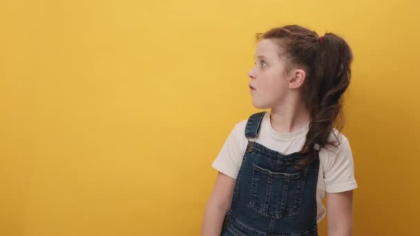 面白い楽しい小さな女の子の肖像画は あなたにカメラを脇に指をポイントし スタジオで黄色の背景色の壁の上に隔離されたポーズ 広告のためのスペースをコピーします 子供のライフスタイルのコンセプト — ストック動画