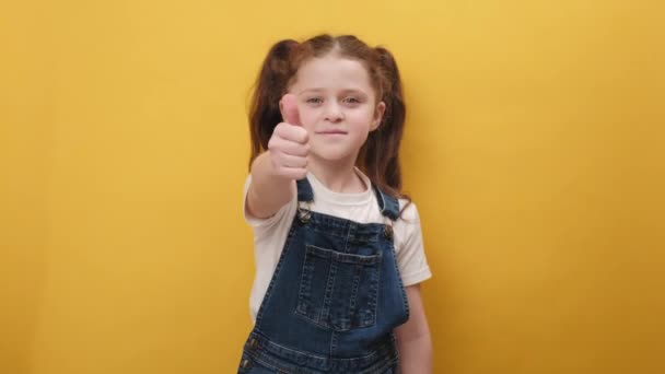 像概念 一个快乐的十几岁以下女孩的画像 她高兴地看着镜头 竖起大拇指 好像是积极的信号 积极的反馈 在黄色的工作室背景下显得孤立无援 — 图库视频影像