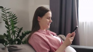 İnternette annelik blogu okuyan, bebeğinin doğum tarihini kontrol eden, online alışveriş sitesinden bebek eşyaları sipariş eden, rahat koltukta oturan cep telefonunu kullanan mutlu genç bir kadının yan görüntüsü.