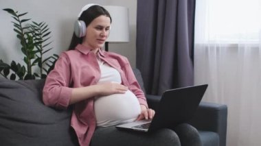 Kulaklıklı mutlu hamile kadın kanepeye oturmuş dizüstü bilgisayarla ailesiyle görüntülü konuşma yapıyor. Müstakbel anne, kadın doğum uzmanı ve jinekologla konuşup profesyonel tıbbi danışmanlık alacak.