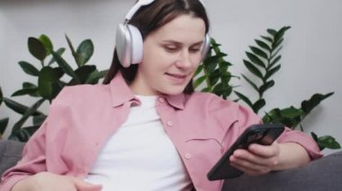 Evde rahat bir koltukta oturmuş kulaklıkla müzik dinleyen mutlu, genç ve hamile bir kadın hamilelik süresince rahatlama ve kişisel bakım, akıllı telefon ve teknoloji, hamilelik ve boş zaman.