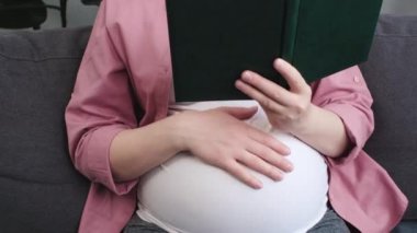 Sevimli, genç, beyaz, hamile bir kadının etrafını sarın. Kablosuz kulaklıkla müzik dinleyip doğmamış çocuğa masal okuyun. Müstakbel anne evdeki oturma odasında rahat koltukta oturuyor.
