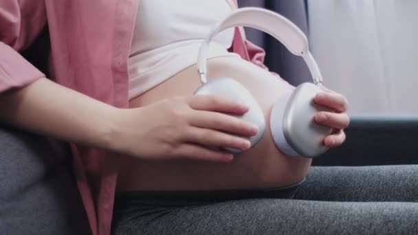 自宅の快適なソファに一人で座って腹にヘッドフォンを適用する若い妊婦のクローズアップ 居心地の良いリビングルームで期待の母親が音楽を聞いている 未来のお母さんが生まれていないリスニング音楽を — ストック動画