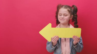 Büyük kağıt ok tutan, kameraya mutlu bakan gülümseyen küçük bir kızın portresi kırmızı stüdyo arka planında reklam için izole edilmiş kot ceket giyiyor. İnsanların duygu kavramı