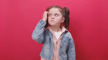 Pensive little kid girl gesturing with raised palms, frowning, being displeased and confused with dumb question, wearing denim jacket, posing isolated over pastel red color background wall in studio