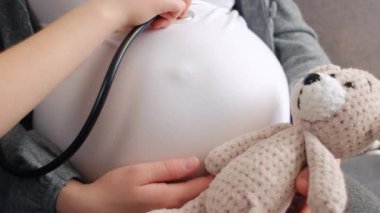 Medikal steteskop kullanan, hamile annesinin koca göbeğini dinleyen, oyuncak ayıcığını kucağında tutan küçük kızı yakından çek. Anne evde çocukla eğleniyor. Çocuk doğumu kavramı