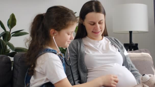 可爱的小女儿用医用听诊器 听怀孕的妈妈大肚子 询问怀孕的事 期待着年幼的弟妹 期待妈妈和十几岁前的女孩坐在沙发上玩乐 — 图库视频影像