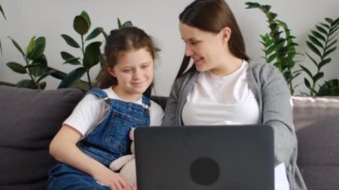 Oturma odasında dizüstü bilgisayar kullanan, internetten film izleyip gülen küçük tatlı kızıyla rahat koltukta oturan mutlu, genç, beyaz bir annenin portresi.