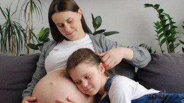 Meraklı küçük kızının sevgi dolu annesinin hamile karnını dinleyip birlikte koltukta oturup gülerken çekilmiş hali. Hamile bir anne, evdeki oturma odasında çocuğu kucaklıyor. Annelik kavramı