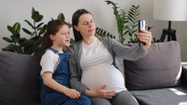 Gülümseyen anne ve kız çocuk resim yapıyor ya da sanal çağrı yapıyor, evde modern akıllı telefondan video bloğu çekiyor. Güzel hamile bir kadın ve güzel bir kız çocuğu telefon kamerasına bakarak selfie çekiyorlar.