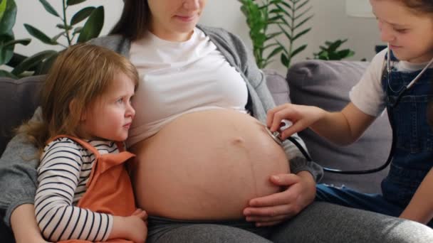 妊娠中の母親の腹に聴診器を適用する愛らしい2人の小さな娘 腹部の弟の動きを聞いて お母さんが子供たちと遊ぶのを楽しみにしています 子供の頃の母親の概念 — ストック動画