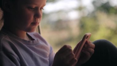 Akıllı telefon kullanarak gülümseyen, internet oyunları oynayan yeşil orman arka planında, pencerenin kenarında oturan güzel küçük bir kız. Yeni nesil ve modern teknoloji aşırı kullanımı