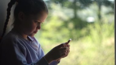 Odaklanmış küçük kız, arka planda güzel bir bahar ya da yaz doğasında pencere kenarına rahatlıyor, cep telefonunda wi-fi kullanıyor, dersten sonra çevrimiçi iletişimin tadını çıkarıyor. Halk yaşam tarzı konsepti