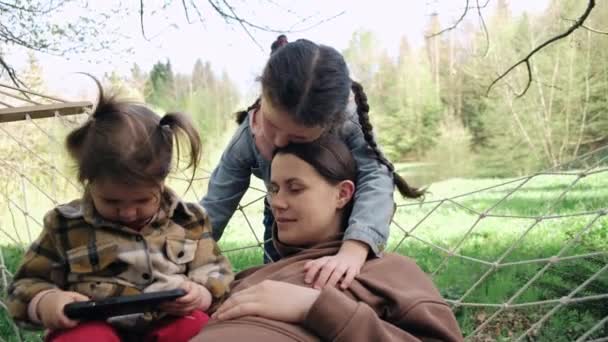 快乐美丽的年轻妈妈和两个可爱的小女儿在吊床里荡秋千 用智能手机 享受周末 在大自然中保持冷静 放松心情 家人在吊床上休息 户外露营生活方式 — 图库视频影像