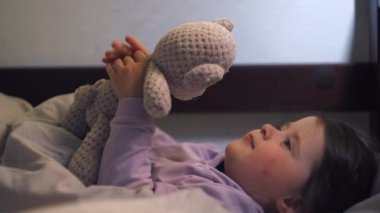 Huzurlu küçük anaokulu kızı oyuncak ayıyla sıcak battaniyenin altında yumuşak yastıkların üzerinde uzanıyor ve iyi geceler diliyor. Mutlu huzurlu küçük kız hafta sonu tembelliğinin tadını çıkarıyor.