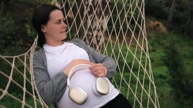 Karnına kulaklık takan çekici ve hamile bir kadının sıcak bir hamakta yatışının yan görüntüsü. Müstakbel mutlu anne Green Park 'ta müzik dinliyor. Hamilelik, annelik ve beklenti konsepti