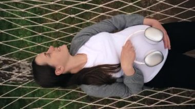 Hamile karnına kulaklık takarak rahat bir hamakta uzanan hamile bir annenin mutlu genç ve mutlu görüntüsü doğmamış bebeğe göbek terapisinde müzik dinletiyor.