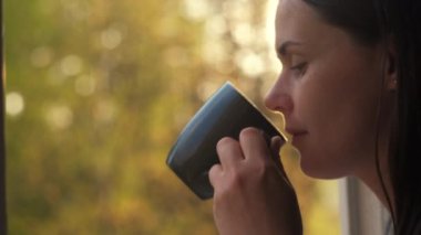 Gülümseyen genç beyaz kadının yakın plan portresi elinde kahve bardağıyla açık otel odasının penceresinin kenarına yaslanmış harika gündoğumu sabahında güzel bir bahar veya yaz ormanı manzarasının keyfini çıkarıyor.