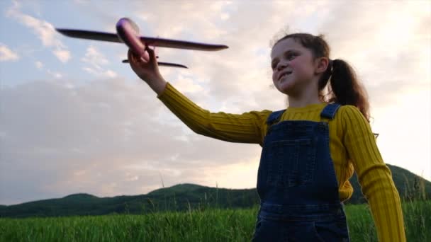 日没の間に背景の空と山でおもちゃの飛行機で遊ぶ遊び心のある女の子のクローズアップ 幼い頃の夢 自由と飛行機の概念 かわいいプリティーチャイルドパイロット スローモーション — ストック動画