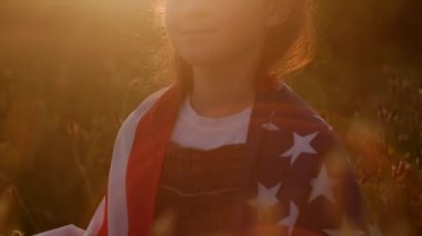 Amerika 'nın 4 Temmuz Bağımsızlık Günü' nde güneşli yaz günbatımında kutlayan tatlı küçük bir kız. Yeşil sahada Amerikan bayrağı taşıyan bir çocuk. Ülke bayrağına sarılmış gururlu küçük Amerikalı çocuk