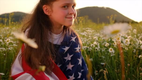 背景にアメリカの小さな子供驚くべき山と夏の夕日 晴れた日の空に対して美しいデイジーフィールドに座っているアメリカ国旗を持つ愛国的な女の子の子供 7月4日のコンセプトを祝う — ストック動画