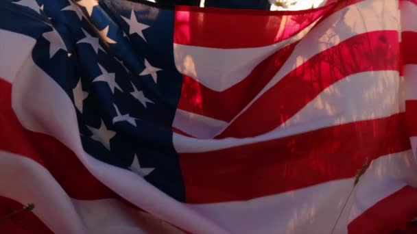 在壮丽的落日下 带着美国国旗在雏菊原野上漫步的小女孩的选择性焦点 美国国旗 爱国主义 独立纪念日7月4日 慢动作 — 图库视频影像