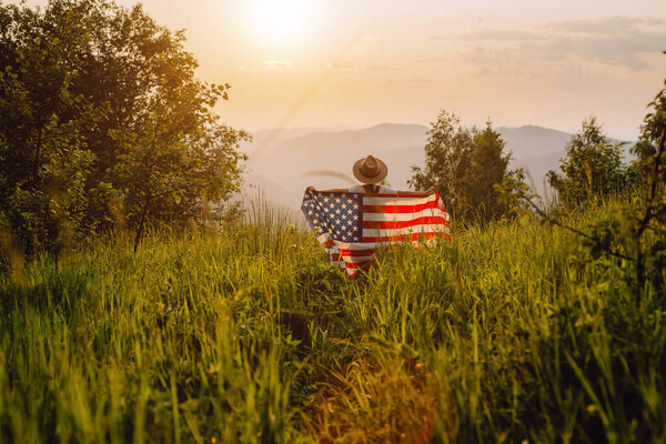 Гордая американка в шляпе, держащая флаг страны. Ребенок с американским флагом США на удивительном фоне гор во время заката. Малыш празднует 4 июля День Независимости США