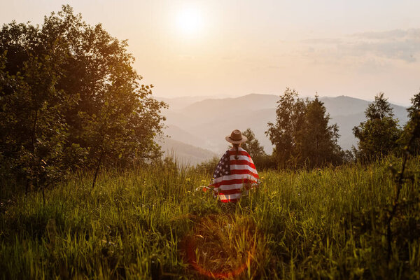 Девочка-подросток в шляпе с американским флагом гордо стоит на фоне гор в золотой час. Концепция Дня памяти, 4 июля, Дня независимости и ветеранов, Празднование США, американских выборов