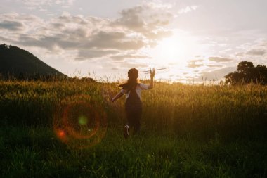 Arka planda uçakla koşan neşeli küçük kız çocuğunun silueti. Muhteşem yaz günbatımı ve görkemli dağlar. Rüya özgürlüğü kavramı. Çocuk el ele tutuşarak sahada koşuyor.