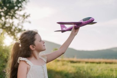 Mutlu küçük kız çocuk arka planda uçak tutuyor yaz günbatımında inanılmaz dağlar. Rüya özgürlüğü kavramı. Oyuncak uçaklı çocuk, güneşli bir günde güzel doğanın tadını çıkar.