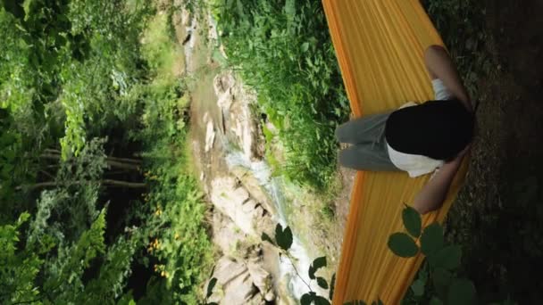 Pionowe Wideo Zrelaksowanej Nastolatki Leżącej Żółtym Hamaku Patrzącej Górską Rzekę — Wideo stockowe