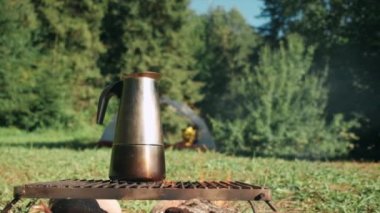 Sıcak bir yaz sabahı kamp ateşinde gayzer kahve makinesinde hazırlanan kahveyi yakın çekim yapın. Yürüyüş yaparken kahvaltı içkisi. Arka plandaki çadır ve ormanda şenlik ateşi. Açık havada aktif macera tatili kavramı