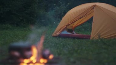 Ormandaki sarı çadırın seçici odak noktası. Sıcak yaz gecelerinde şenlik ateşinde kızarmış sosis ve kaynar su. Hızlı yemek kampı. Ateşte sosisli sandviç. Kamp ateşinde ızgara sosis.