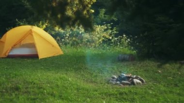 Turist sarı çadırı ve güzel çayırların yanındaki yeşil çayırda kamp ateşi. Yaz kampında doğa arka planında kamp yeri. Macera, seyahat, aktif yaşam tarzı, özgürlük ve açık hava konsepti