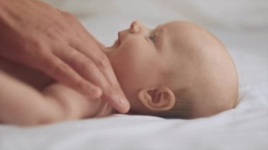 Bakımlı genç bir babanın, rahat bir yatakta uzanan sağlıklı bir bebeğe masaj yapması. Babası karnına masaj yaparken ve kaslarını geliştirirken küçük beyaz bir çocuk sırt üstü uzanıyor.