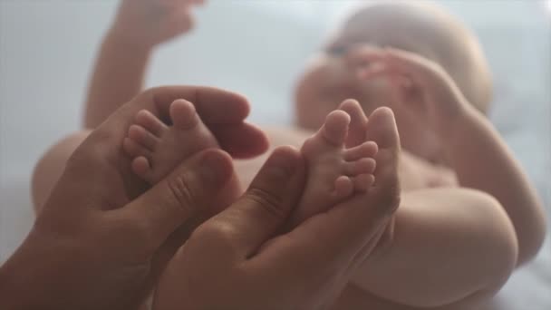自宅で快適なホワイトベッドに横たわる赤ちゃんマッサージを作る若い父親の選択的な焦点 愛する父親は裸足をマッサージし 新生児のための予防マッサージ 両手で赤ちゃんの足をストローキングする父親 — ストック動画