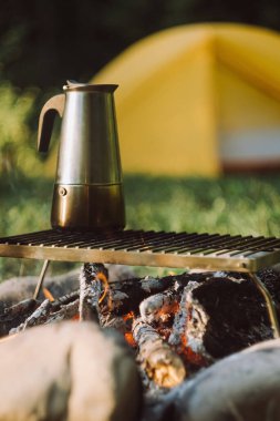 Güneş doğarken ya da batarken yeşil yaz parkında çadırın yanındaki şenlik ateşinin üzerinde gayzer kahve makinesinin yakın çekimi. Kamp ortamı, doğa ve açık hava yaşam tarzı konsepti. Yazın kamp ateşi, sıcak kahve.