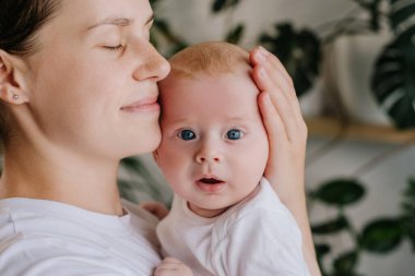 Şefkatli anne kucaklar küçük bir çocuğu okşar. Sevgi dolu genç annenin yakın plan portresi kucak kucak kucak kucak kucak kucak kucak kucak küçük bir çocuk oturma odasında ev bitkileriyle duruyor. Annelik kavramı