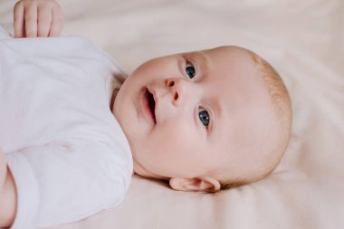 Beyaz elbiseli mutlu erkek bebek portresi evdeki rahat yatakta yatıyor. Sevimli küçük bebek gülümsüyor ve çarşaflarda dinleniyor. Yeni doğan beyaz bebeğin yatak odasındaki üst görüntüsünü kapat. Çocukluk kavramı