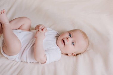 Beyaz tulumlu tatlı bir bebeğin portresi evde rahat bir yatakta uzanıyor, gülüyor, kameraya bakıyor. Gülümseyen güzel bebeğe yakın dur. Bebeklik, sağlık, pediatri, çocuk bakımı konsepti