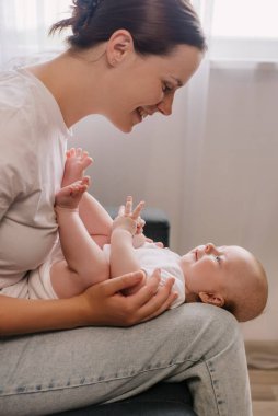 Üç aylık bebeğiyle evdeki rahat koltukta oynayıp anneliğin ve doğum izninin tadını çıkaran mutlu bir anne. Sevimli bebek kucak annesi oturma odasında uzanıyor, gülüyor.