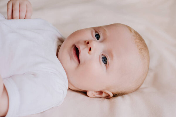 Портрет счастливого мальчика в белом боди-костюме, лежащего на уютной кровати дома. Симпатичный малыш улыбается и расслабляется на простынях. Закройте вид сверху на новорожденного ребенка в спальне. Концепция детства