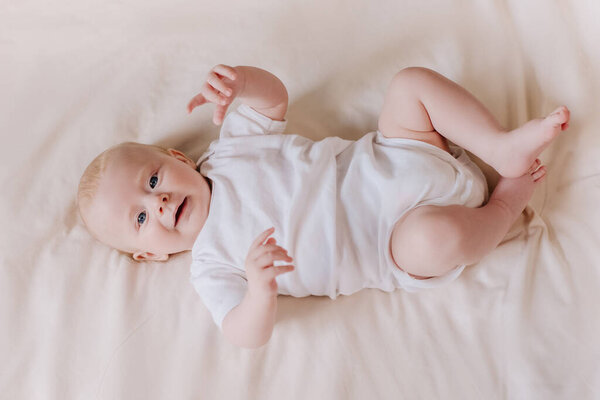 Top view of happy adorable little newborn boy with smiling face lying on cozy bed in living room at home. Cheerful pretty infant baby resting playing lying down on comfy blanket. Childhood concept