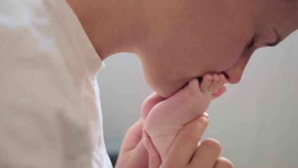 专心致志的年轻母亲亲吻坐在舒适的沙发上的可爱新生儿的小脚 对婴儿 快乐的母亲 儿童保育的理念表达真诚而又温柔的感激之情 — 图库视频影像