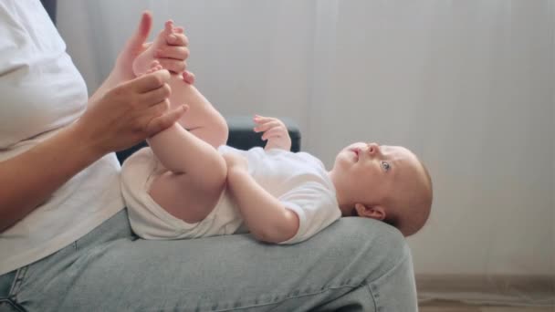 快乐的年轻可爱的妈妈轻轻地吻着坐在舒适沙发上的小脚 穿着紧身衣的新生儿的小脚趾 与孩子 母亲和后代的温馨时刻 母亲关心的概念 — 图库视频影像