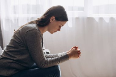 Evdeki pencerenin yanındaki kanepede oturan mutsuz genç kadın, sorun ve sorunları düşünüyor, depresyon ya da yalnızlık çeken üzgün kadın, zihinsel hastalık, zor kararlar veriyor.