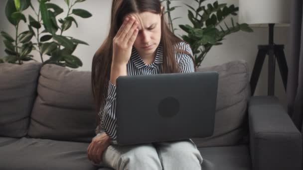 疲れている悲しい若い女性は ノートパソコンでソファーに座っている問題を心配し オンラインで悪いニュースを読んだり 借金や否定的なメッセージに関する電子メール通知に悩まされた女性を強調しました — ストック動画