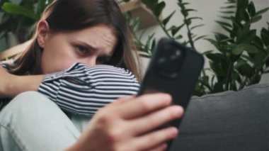 Evdeki kanepede oturan düşünceli, genç, beyaz bir kadının cep telefonuyla sorunlarını düşünüp durduğu yakın plan. Gergin esmer kız, akıllı telefondaki mesaj ya da mesajdan rahatsız oluyor.