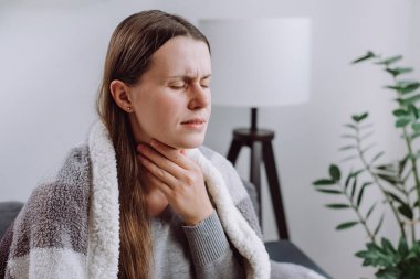 Symptoms from flu season in winter concept. Close-up of unhealthy sad young woman covered blanket sitting on grey couch at home suffering from sore throat, angina, hard to swallow, voice loss clipart